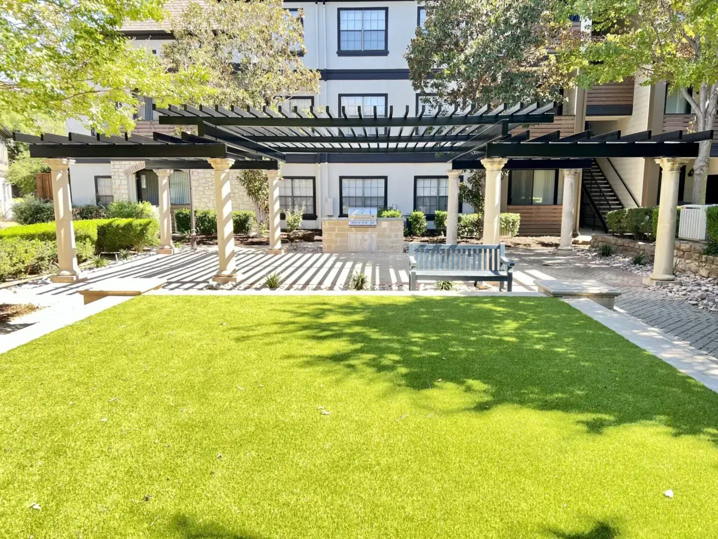 Outdoor patio with grill, pergola, bench, and artificial lawn in front of an apartment building.