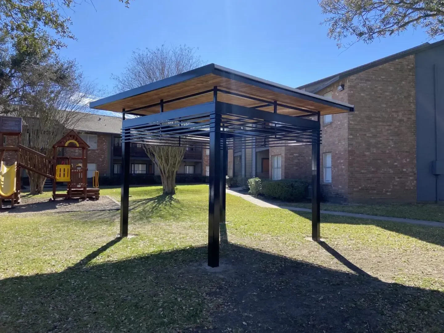 Black pergola with a wooden roof over a grassy area, in front of an apartment building on a sunny day.