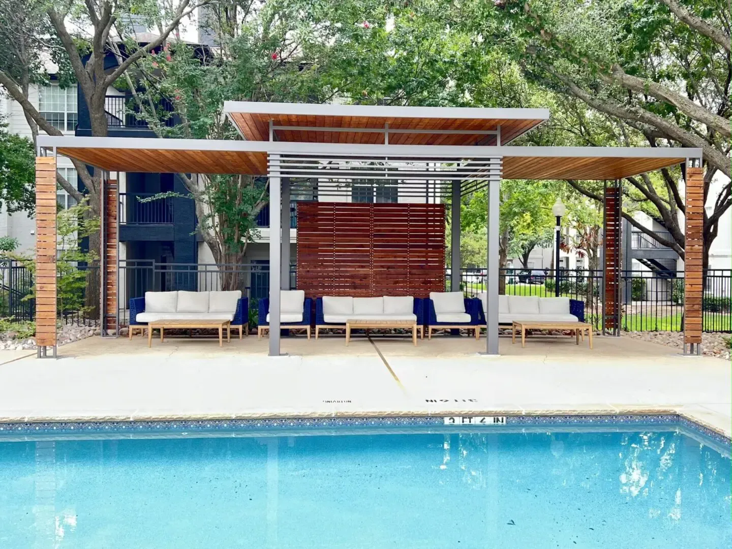 Poolside lounge area with couches under a wooden pergola. Blue pool water.