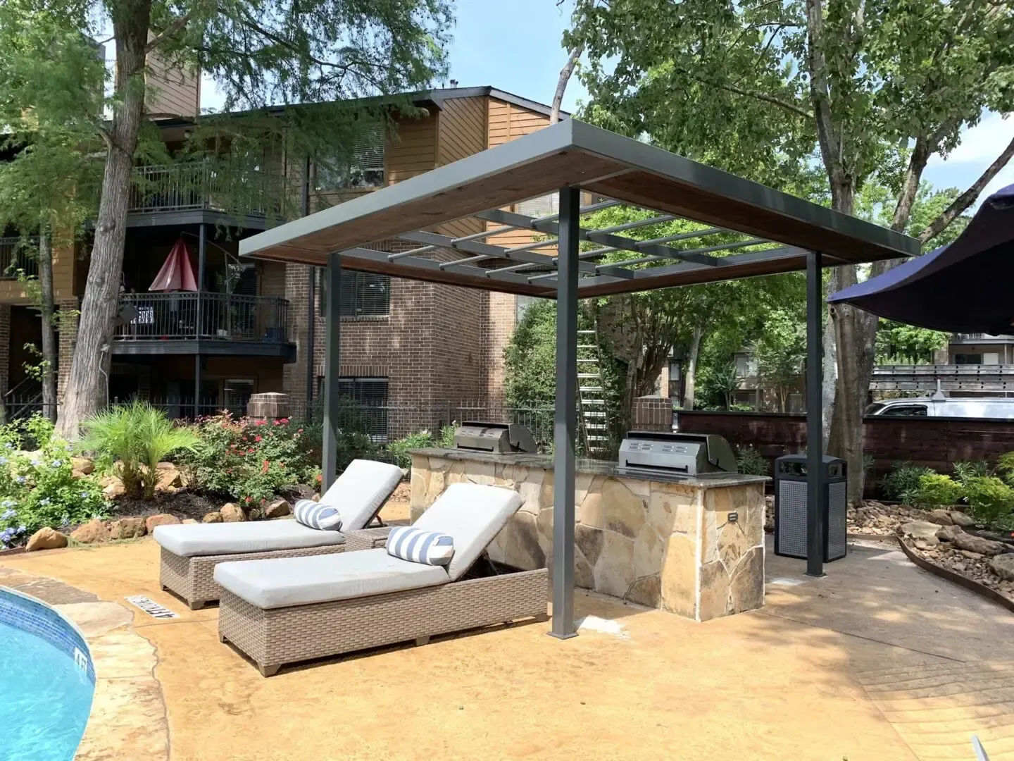 Poolside shaded cooking area with lounge chairs, stone counter, grill, and trash can.