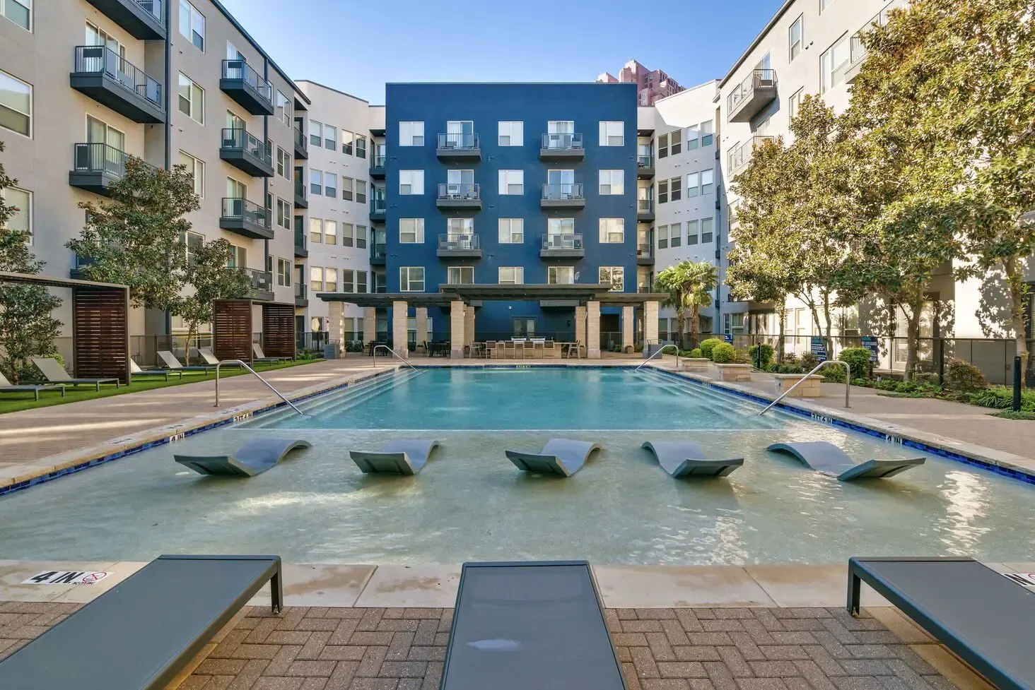 Swimming pool in a modern apartment complex. Lounge chairs in water and on deck. Blue building backdrop.