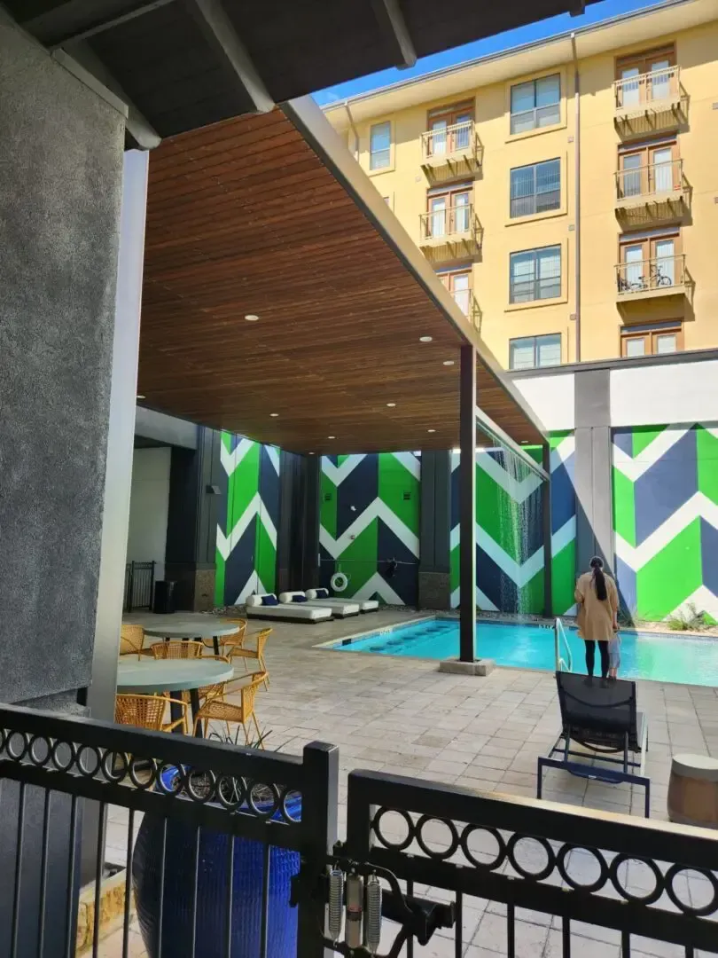 Poolside patio with tables, pool, and person; building in background; decorative green and blue chevron wall.
