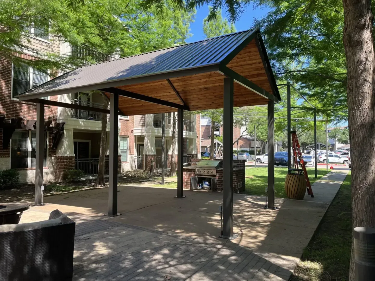 Outdoor BBQ area with a metal-roofed shelter, brick grill, and nearby apartment buildings.