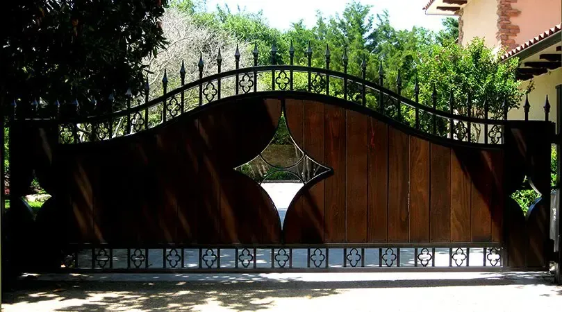 Wooden driveway gate with diamond cutouts, topped with wrought iron, set against a green foliage backdrop.