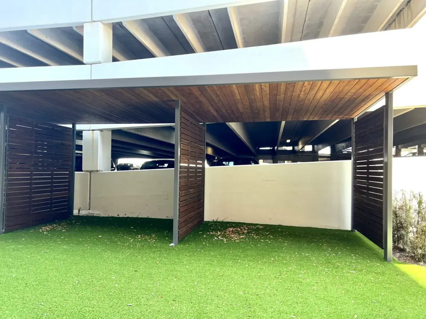 Car shelter with wooden accents, green turf, under a parking garage.