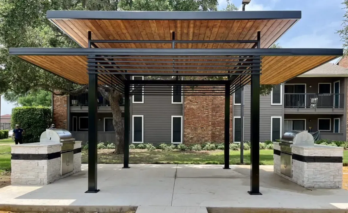 Gazebo with black metal frame, wooden ceiling, and stone grill stations in front of a brown apartment building.