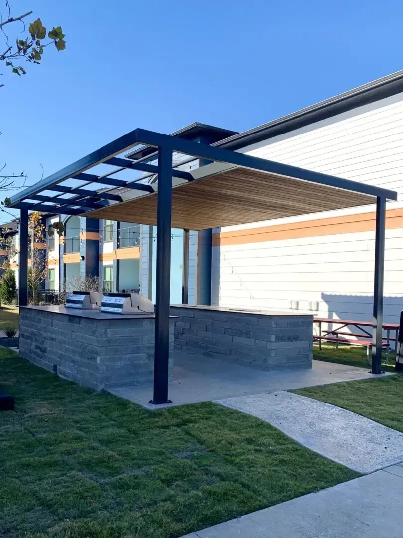 Pergola over a stone counter, next to a building and grass.