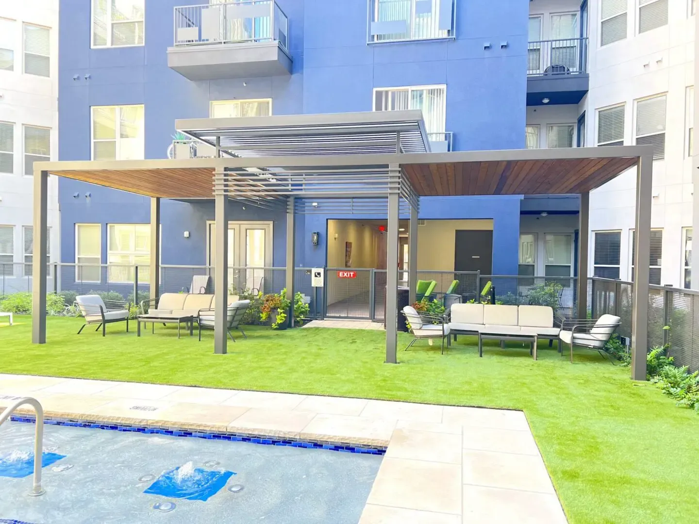Outdoor seating area with sofas, chairs, and pergola by a pool, in front of a blue building.