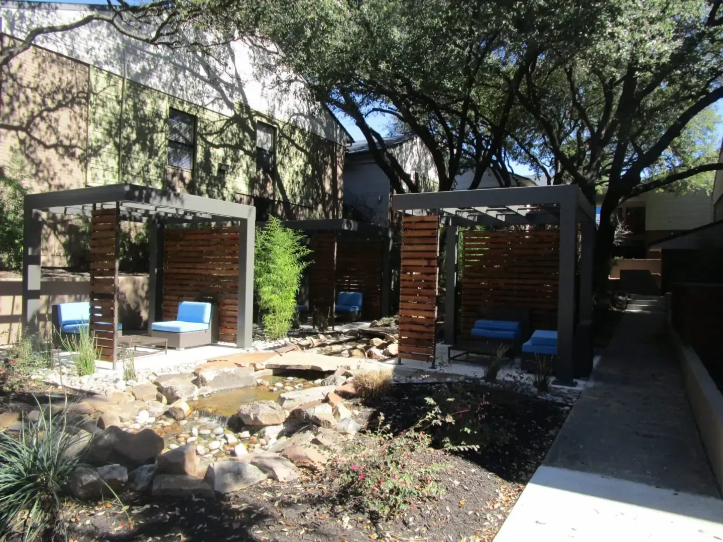 Outdoor seating area with wooden structures, blue chairs, small water feature, and lush greenery.