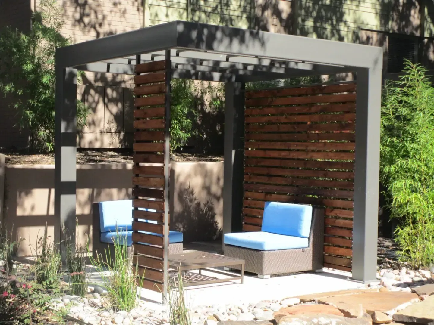 Modern outdoor seating area with grey pergola, wooden privacy screens, blue cushions, and plants.