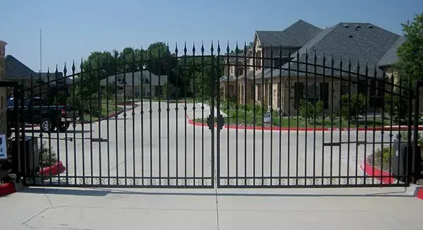 Black wrought-iron entrance gates to a gated community on a sunny day. Houses visible in the background.