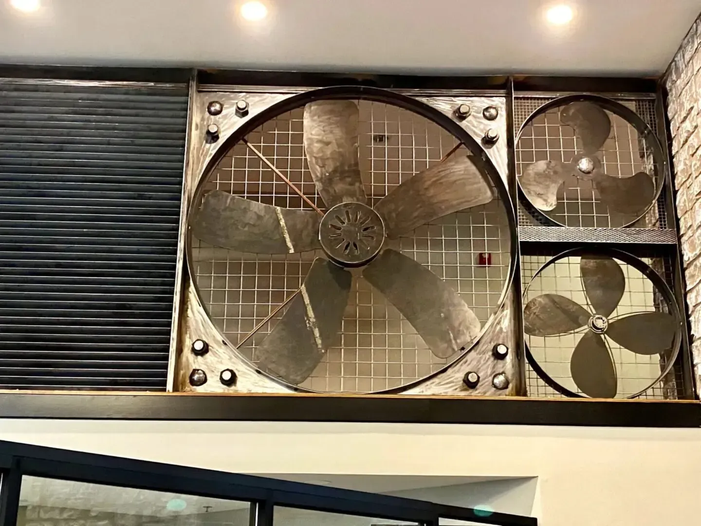 Industrial-style fans, varying sizes, mounted on a wall. Next to a black shutter. Metallic and brick setting.