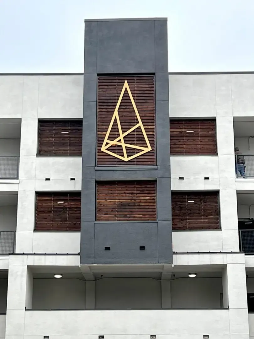 Modern building facade with a dark gray central tower with wood accents and a gold geometric logo.