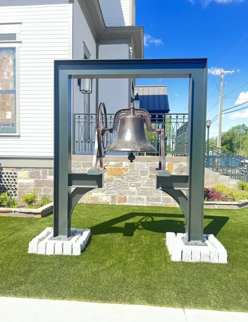 Bronze bell in a dark metal frame on a brick base, standing on green grass, with a modern building in the background.