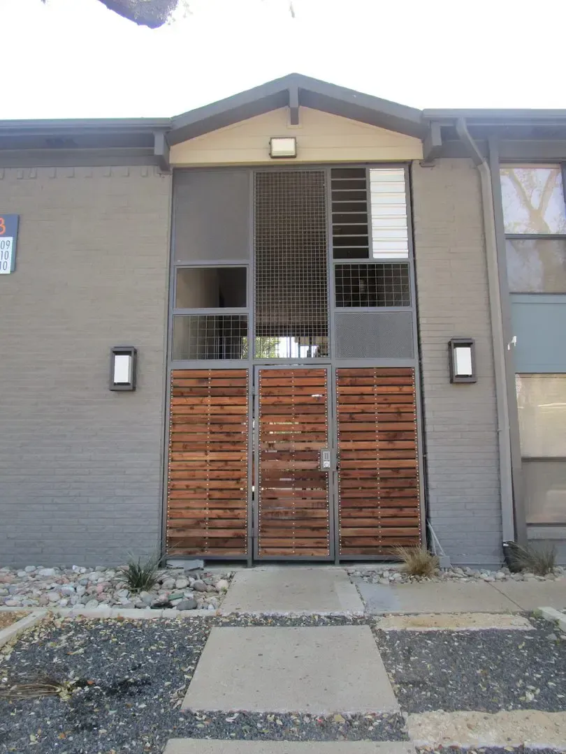 Apartment entrance with wooden door, metal screen, and gray exterior. Pathway leads to the door.