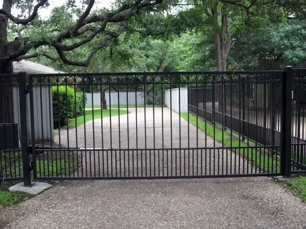 Black metal driveway gate on a concrete and gravel surface; lush green trees in the background.