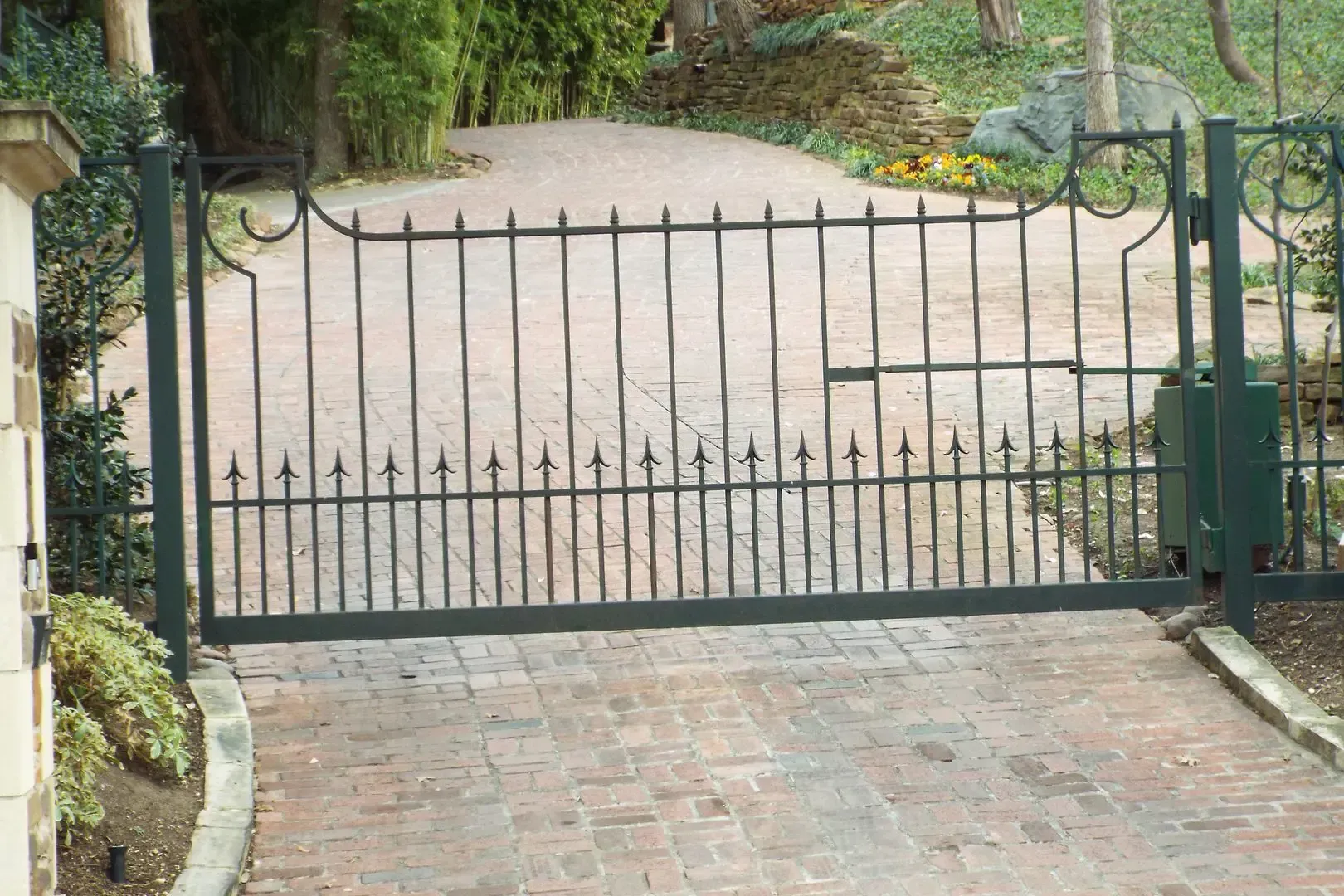 Black wrought iron gate across a brick driveway leading to a tree-lined property.
