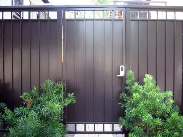 Black metal gate with a lock, flanked by green bushes.