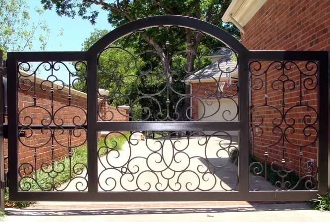 Ornate wrought iron gate, arched top, intricate scrollwork, brick house background.