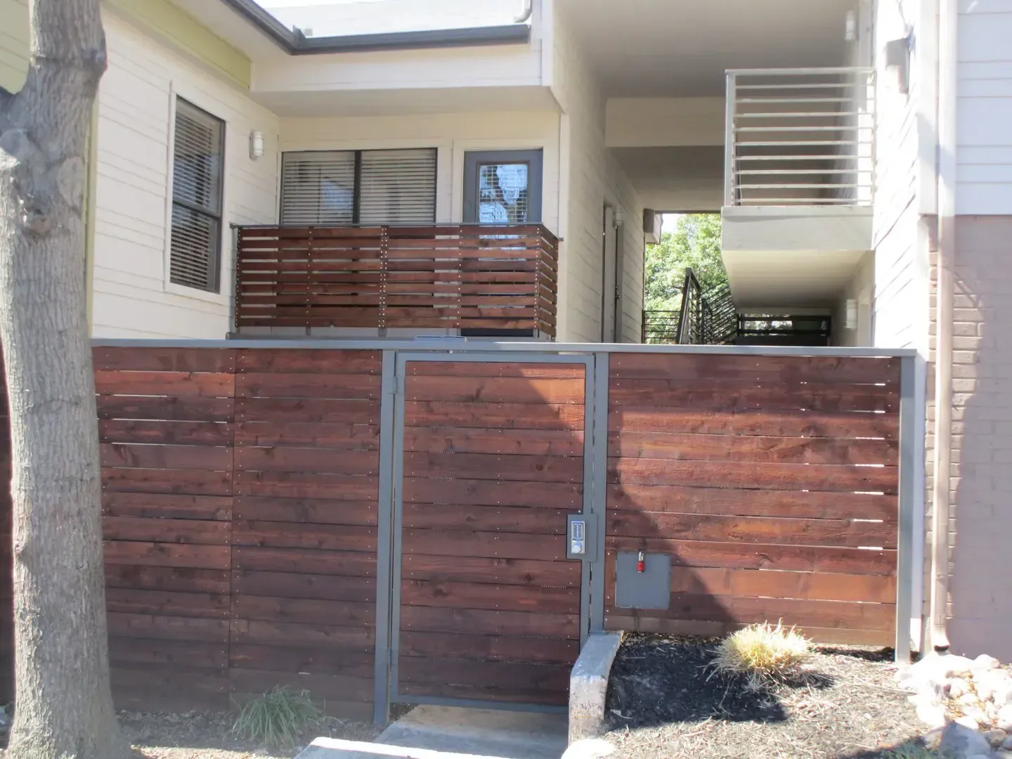 Wooden fence and gate in front of a building with a balcony. Dark wood, gray frame, green trees.