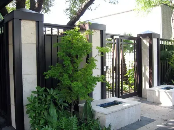 Black and stone fence with gate, lush green plants, and square stone planters.