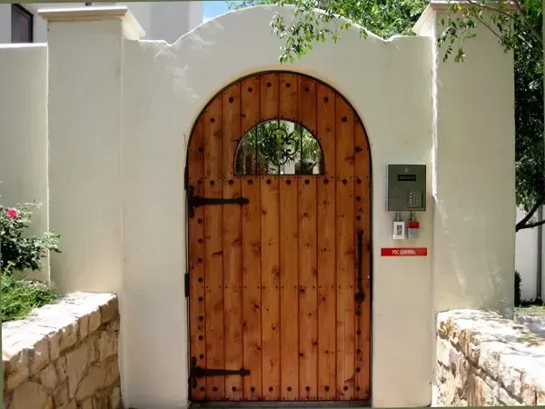 Wooden arched gate in cream-colored stucco wall with a mailbox and alarm on the side.