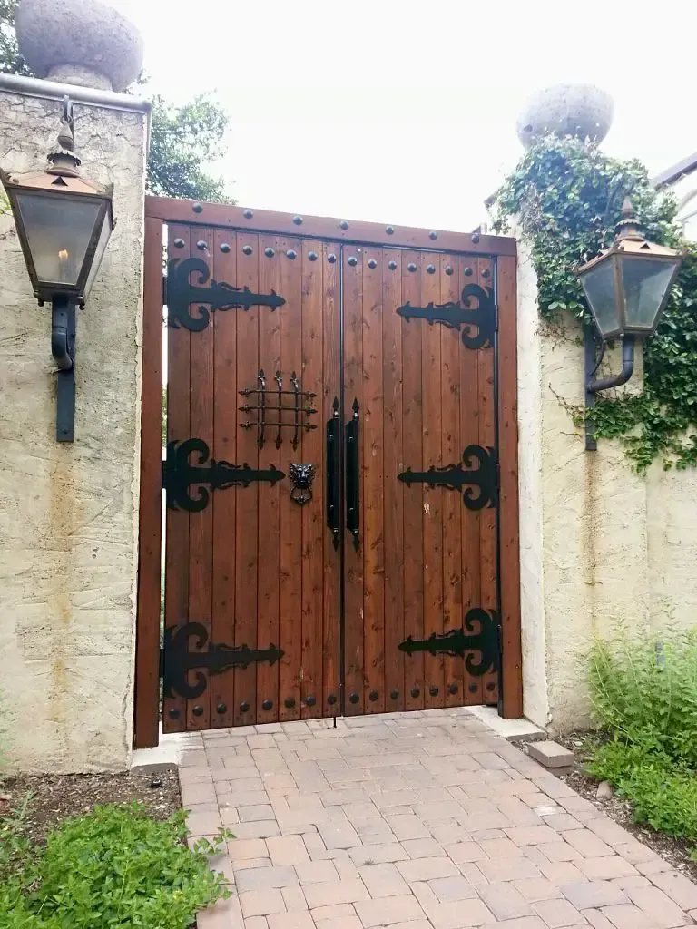 Wooden gate with ornate black hardware, between stucco walls with lanterns. Brick path leads to it.