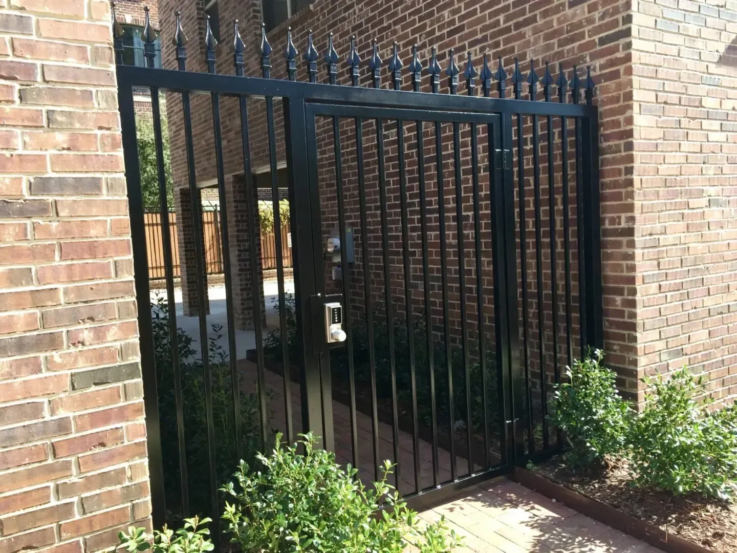 Black metal gate with vertical bars and spikes, between brick walls, secured with a lock.