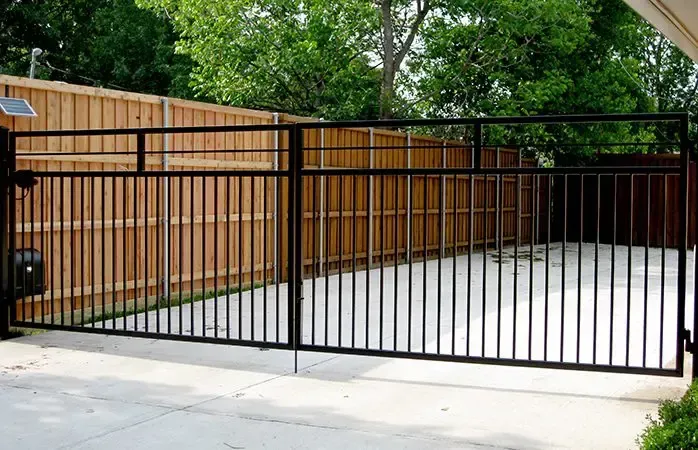 Black metal driveway gate in front of a wooden fence. The ground is concrete with a lawn and trees in the background.