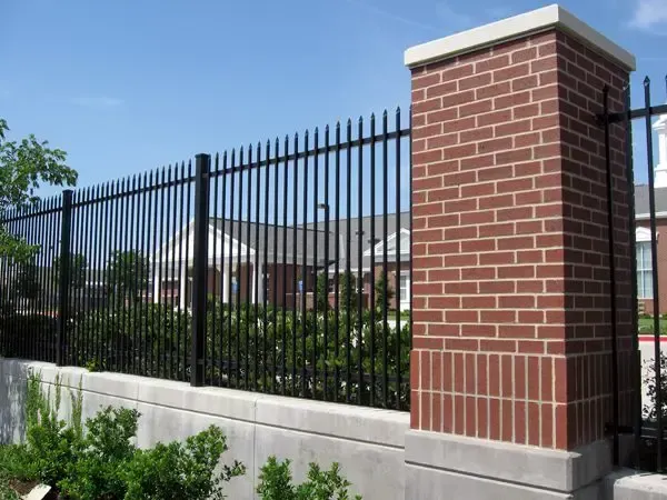 Black metal fence atop a concrete wall with brick pillars; green foliage in foreground.