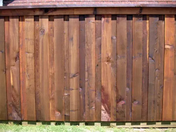 Wooden fence, stained reddish-brown, with vertical planks. Background of green grass and a roof.