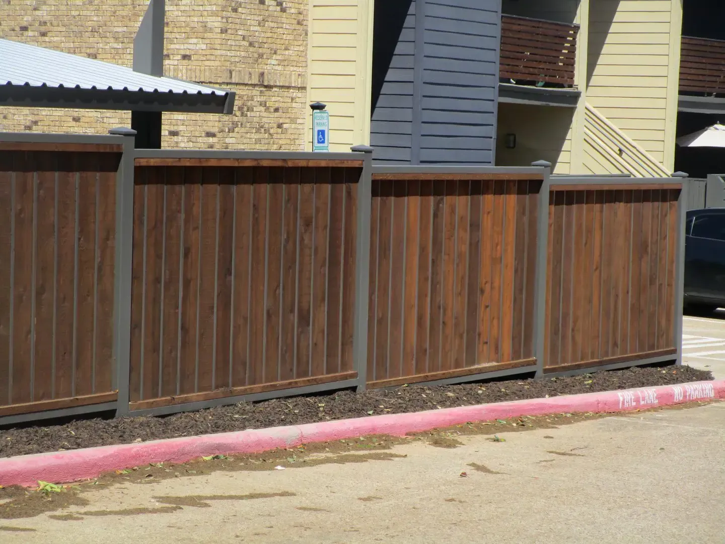 Wooden fence with dark stain, bordered by dark metal posts, next to tan and brown building.