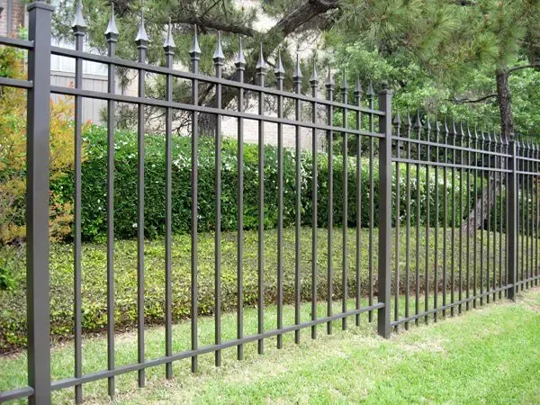 Black metal fence with pointed tops on green lawn, separating the yard from shrubbery.