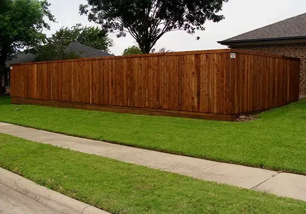Brown wooden privacy fence in front of green grass and a house, with a sidewalk in view.