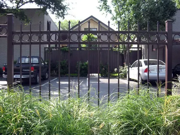 Iron fence in front of a parking area. Cars are parked inside; green bushes in foreground, building visible.