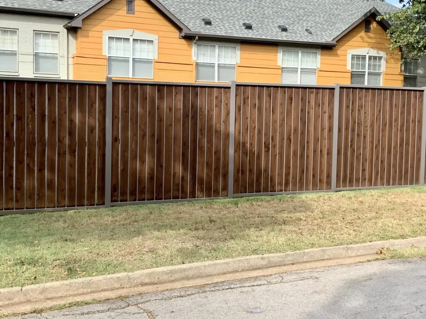 Brown wooden fence in front of a yellow house, with green grass and a paved road in the foreground.