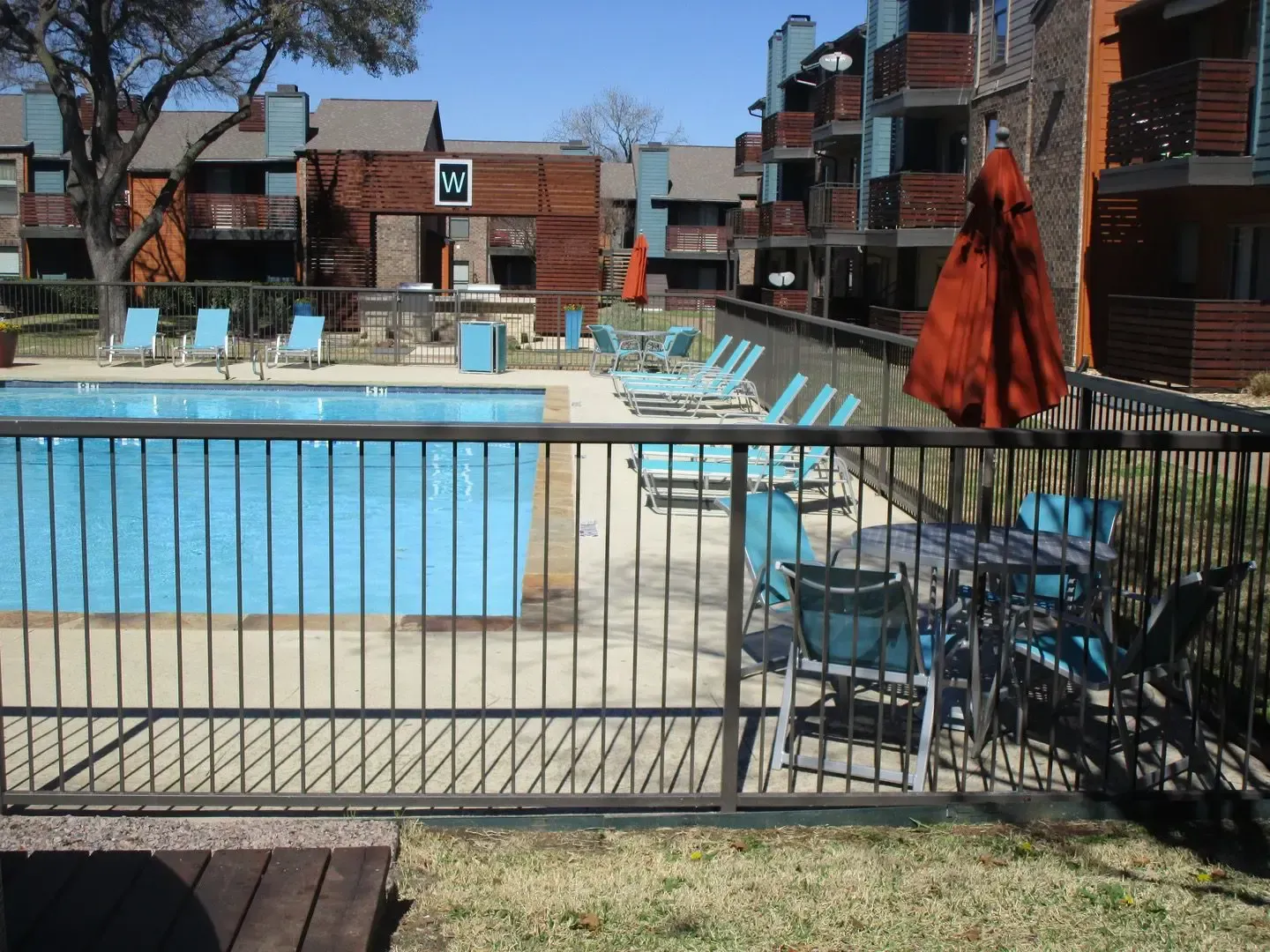 Pool area with blue water, lounge chairs, and a rust-colored umbrella. Buildings in the background.