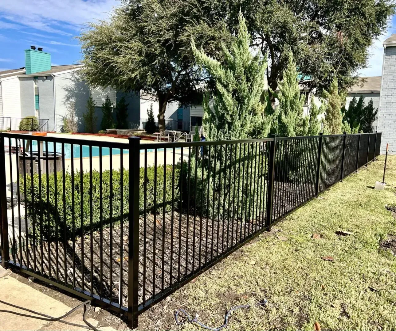 Black metal fence around a pool and bushes on a grassy area. Apartment buildings in the background.