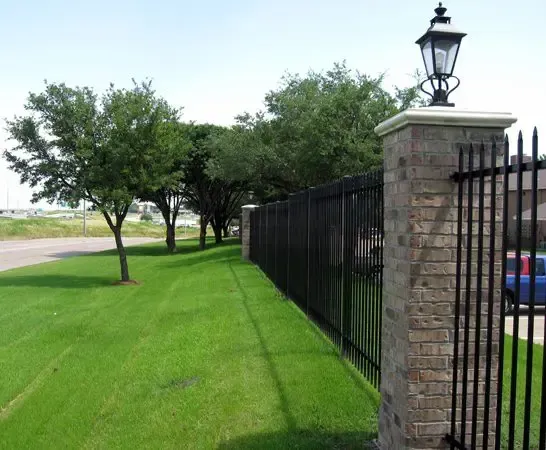 Lush green lawn beside a black metal fence with brick columns and a street lamp.
