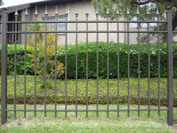 Metal fence with vertical bars and decorative top, fronting a hedge and house.