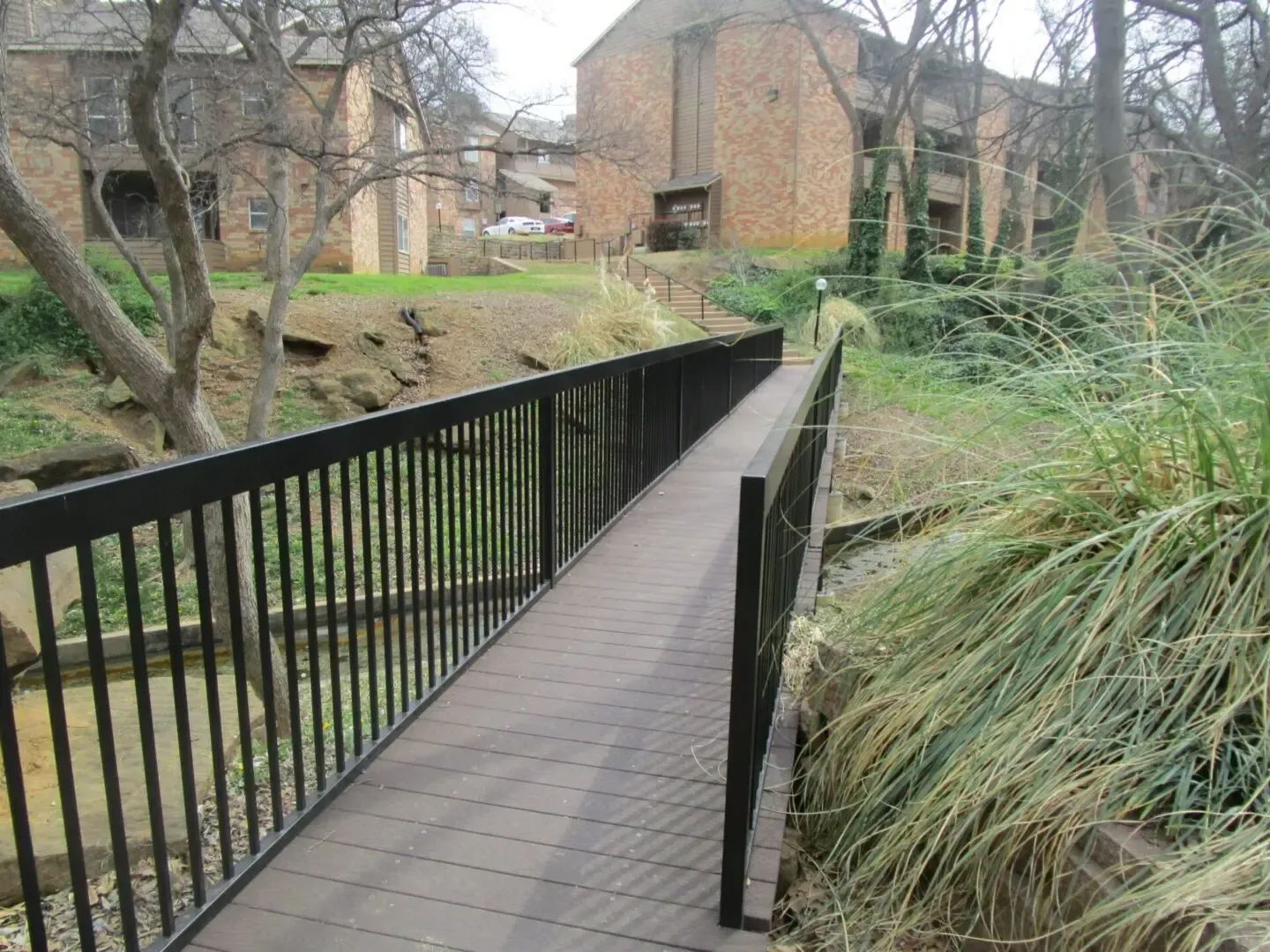 Wooden bridge with black railings over a small stream, leading to apartment buildings.