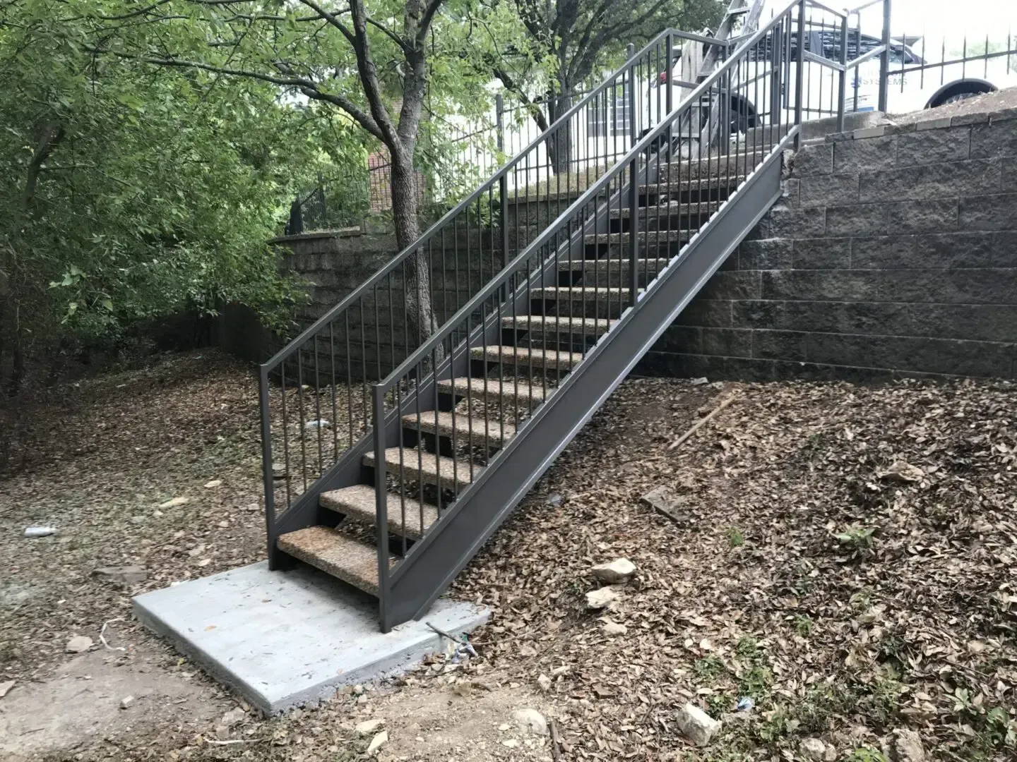 Metal staircase with handrails on a hillside, leading up to an elevated area, surrounded by foliage.