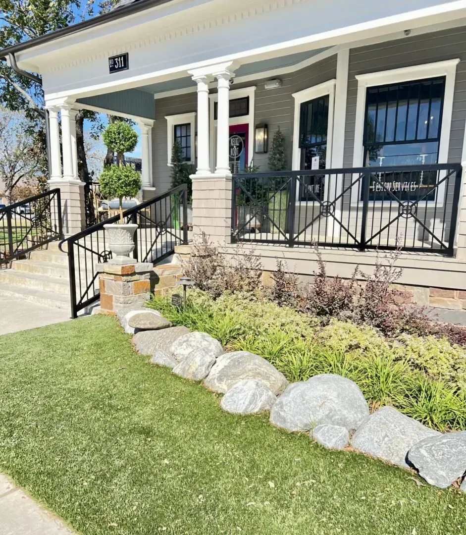 A building with a porch and black railings. Landscaping includes rocks, bushes and green grass.