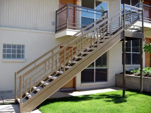 Exterior stairs leading to a balcony. Tan railings and steps, off-white building, green lawn, and sunny setting.