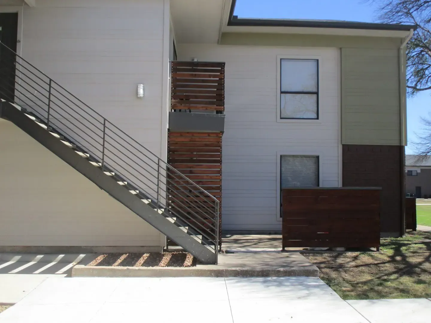 Exterior view of a two-story apartment building with stairs, wooden accents, and a concrete walkway.