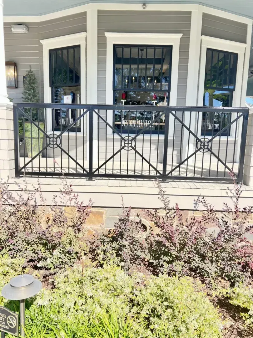 Black iron railing in front of a building with gray siding, windows, and greenery.