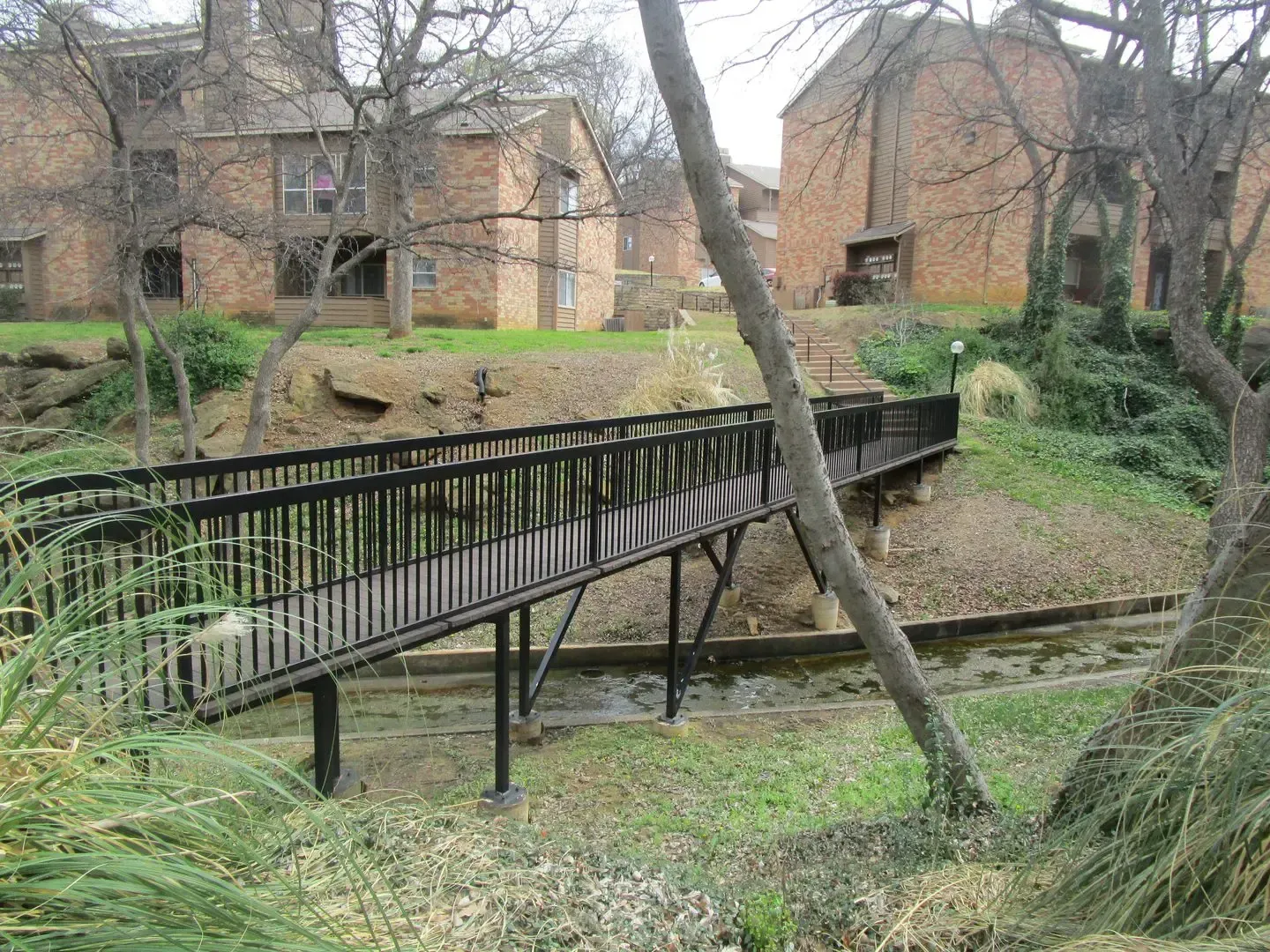 Wooden bridge over a creek in front of brick apartment buildings.