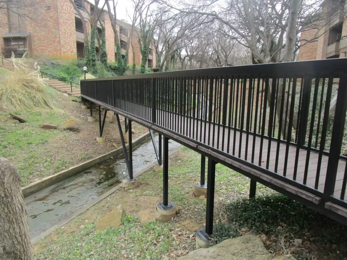 Black metal bridge over a dry creek bed in an outdoor setting. Apartment buildings are visible in the background.