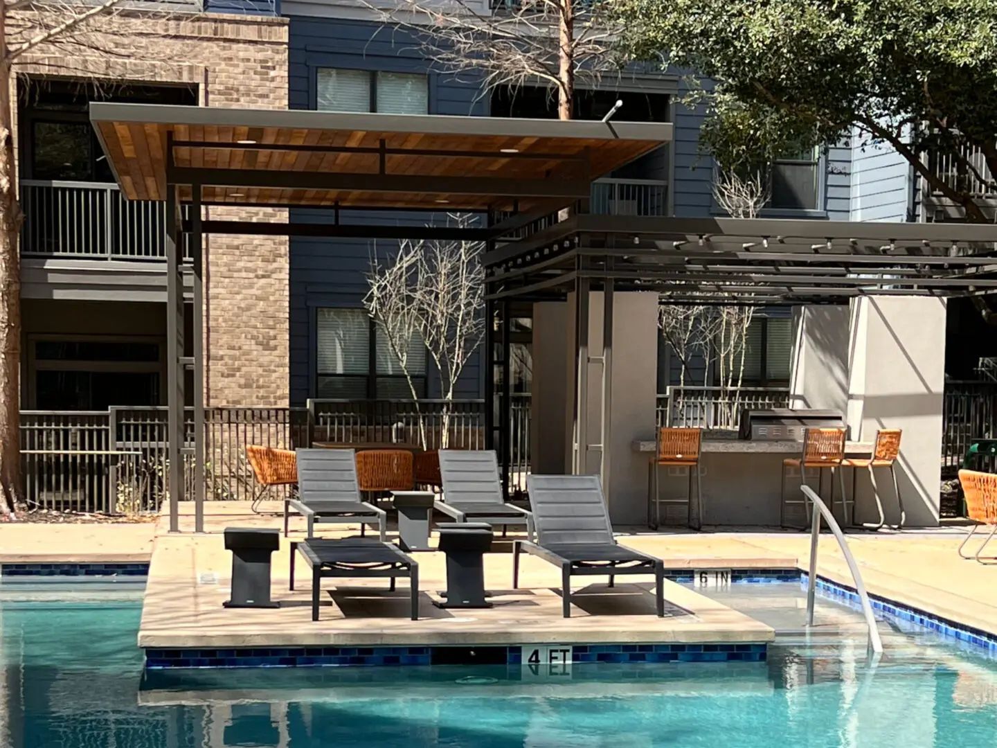 Pool area with lounge chairs under a canopy, bar, and apartment building in the background.