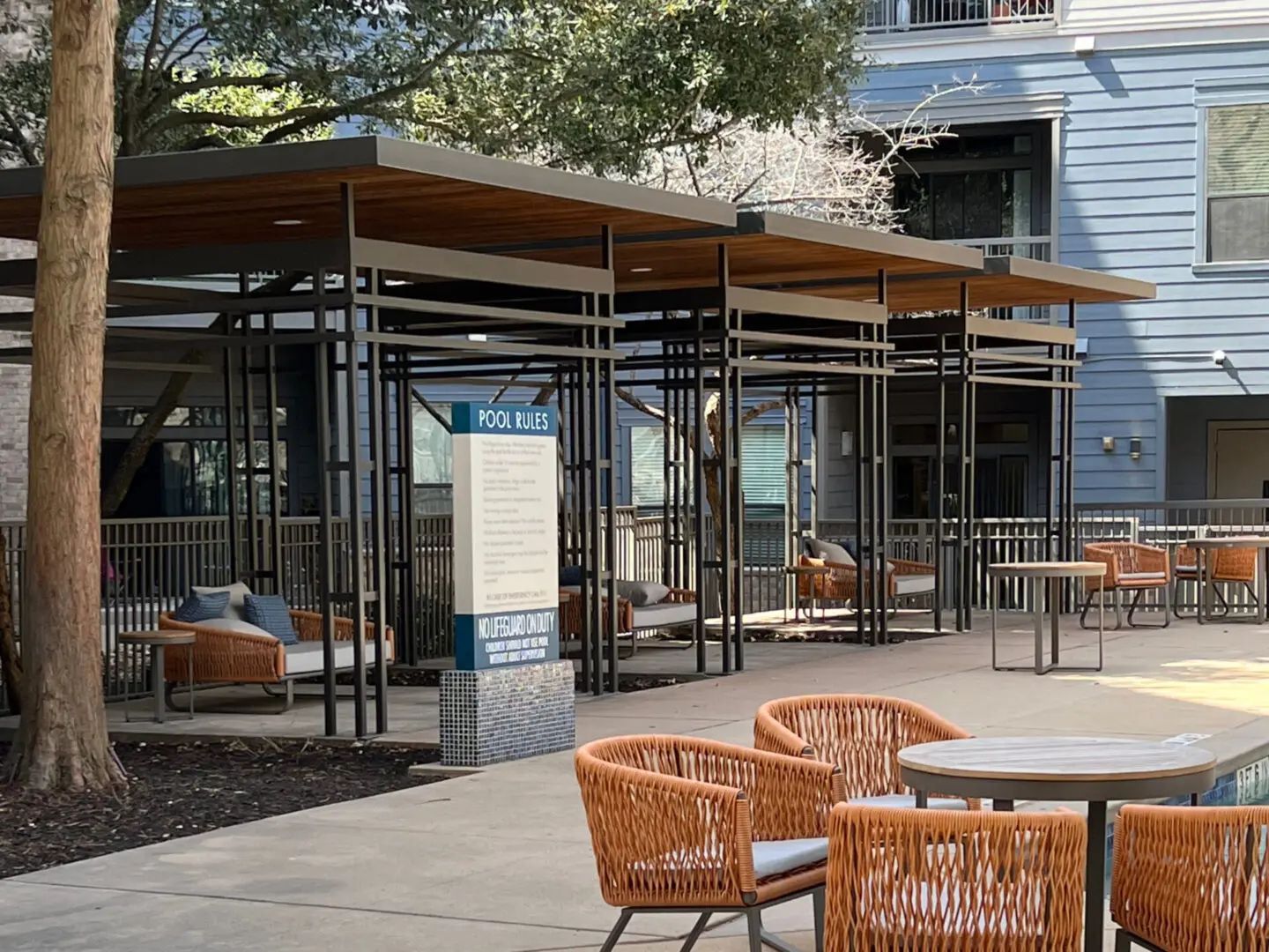 Outdoor seating area with woven chairs and a metal-framed shelter. Blue building in the background.